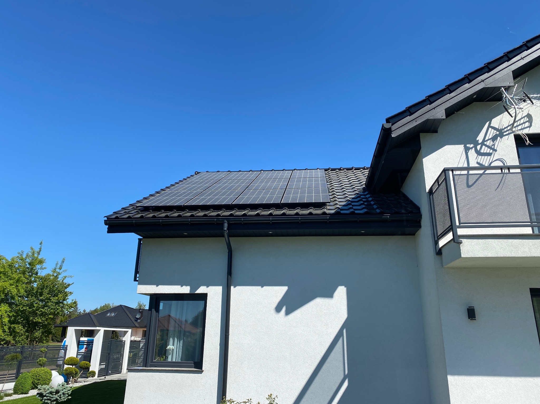 Panele słoneczne na dachu nowoczesnego domu z białą elewacją i czarnymi detalami, błękitne niebo w tle. Widok z dołu podkreśla architekturę i instalację.