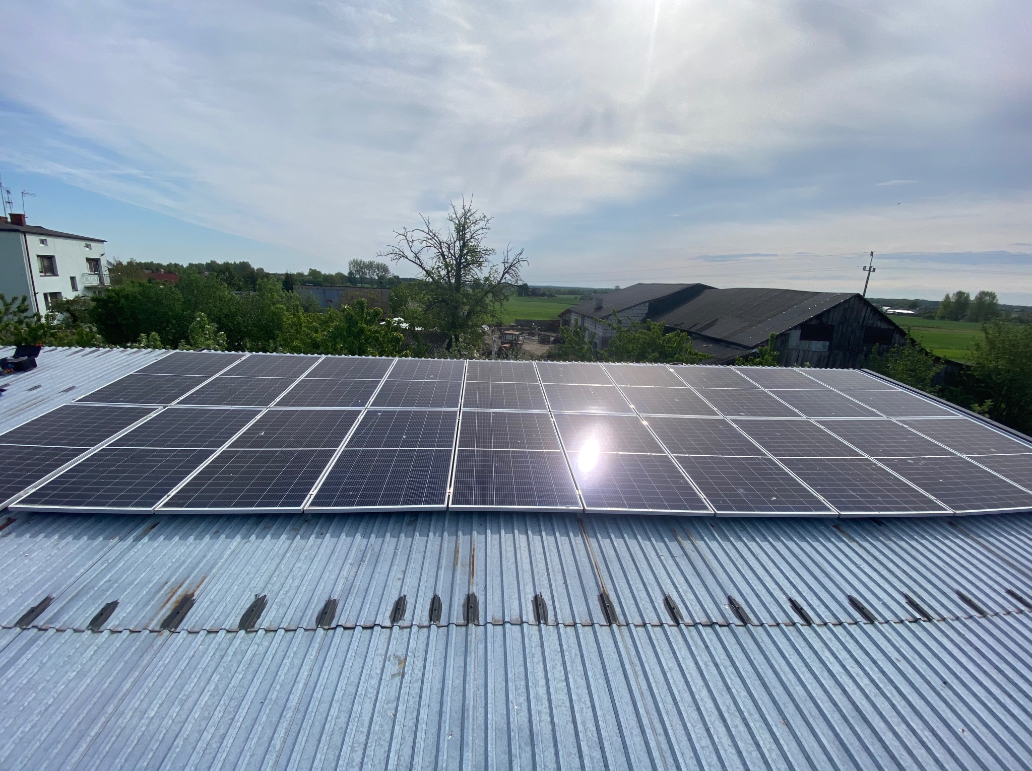 Panel słoneczny na metalowym dachu z widokiem na budynki gospodarcze i drzewa w tle. Jasne niebo z chmurami.