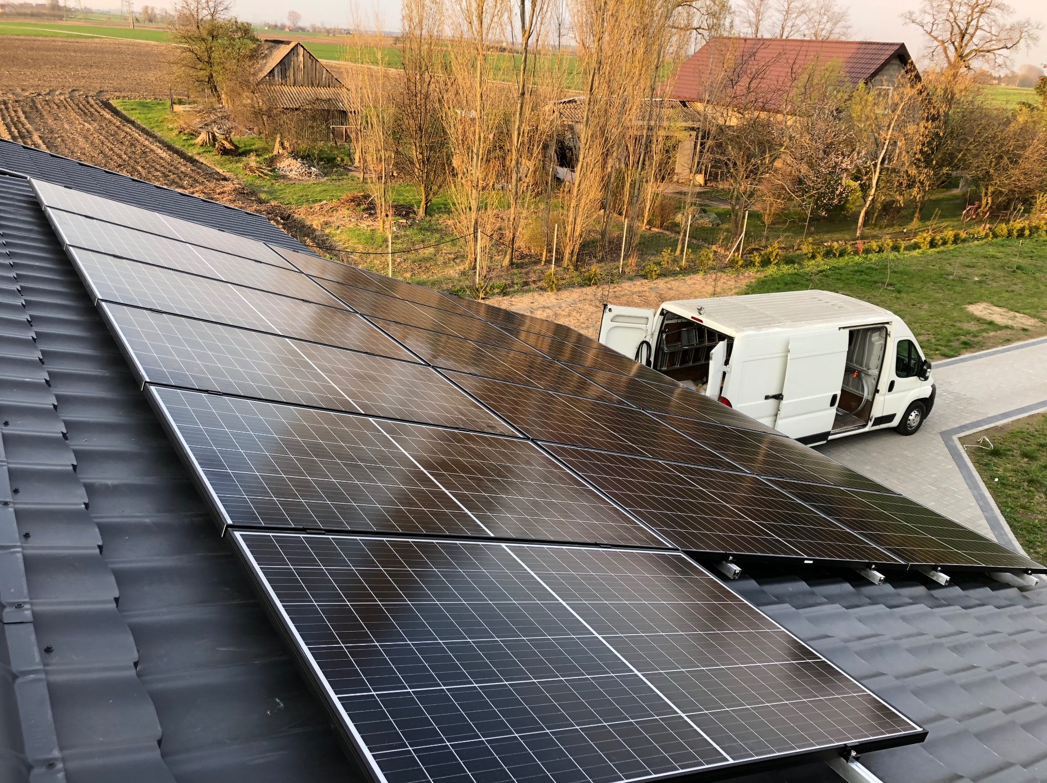 Panele fotowoltaiczne na dachu domu jednorodzinnego, z białym vanem serwisowym w tle i wiejskim krajobrazem, uchwycone w ciepłym świetle słonecznym.