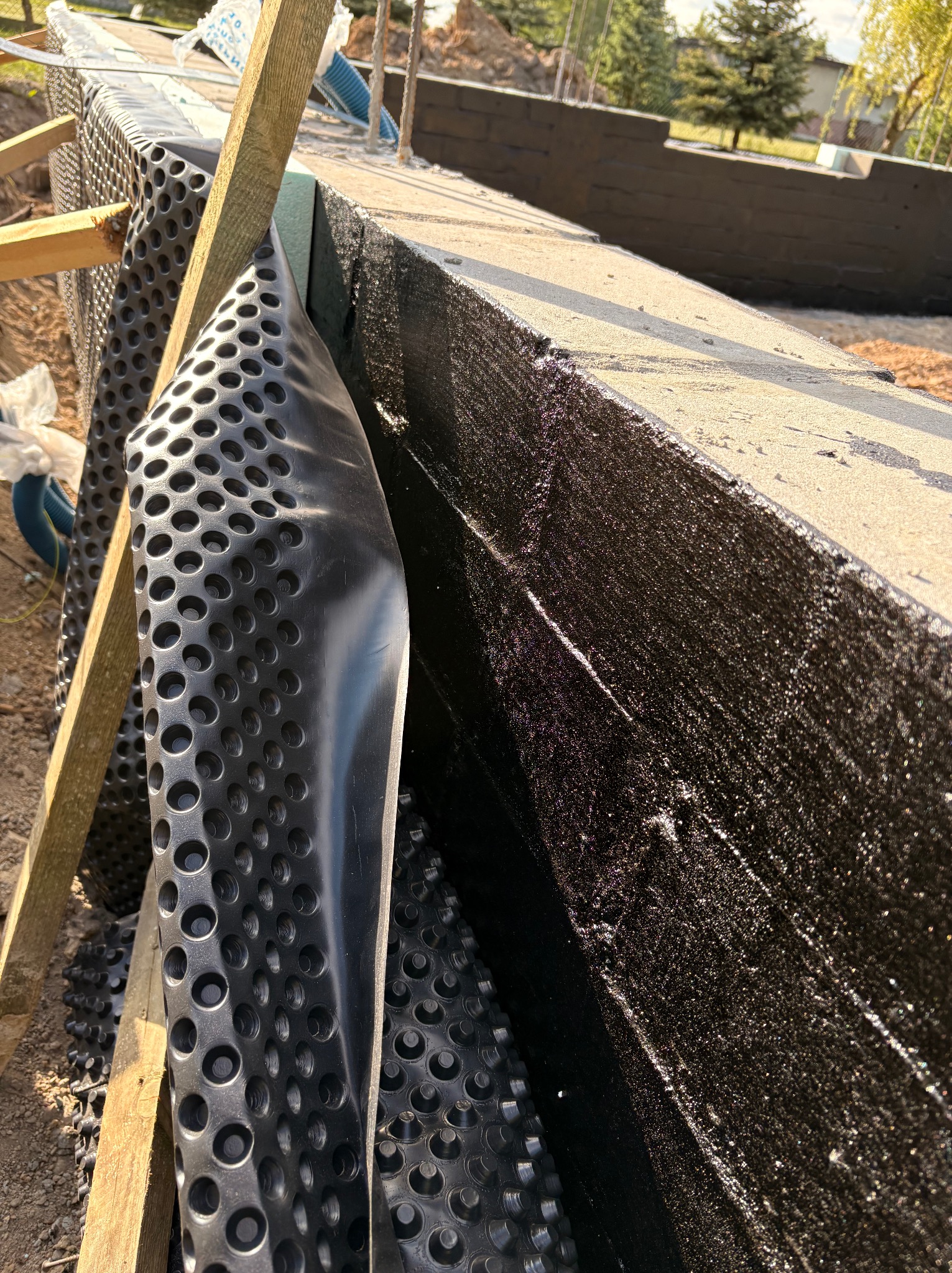 Izolacja fundamentów budynku z użyciem folii kubełkowej i hydroizolacji bitumicznej w słoneczny dzień, widoczne drewniane elementy konstrukcyjne.