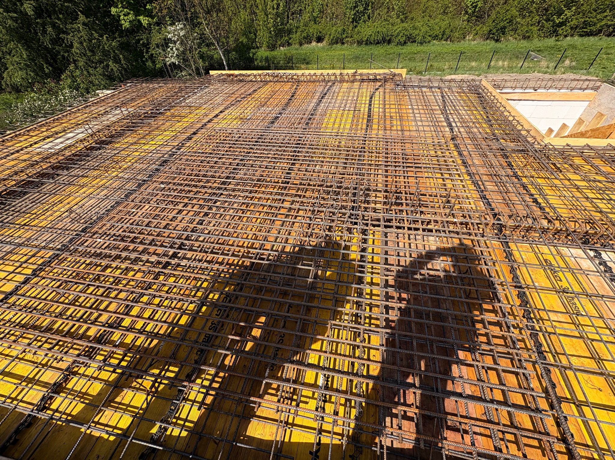 Zbrojenie stropu na żółtym szalunku z widocznym cieniem osoby, przygotowane do wylania betonu, z zielenią w tle. Kąt ujęcia z góry, ukazujący siatkę prętów.