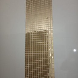 GLAZ-BUD KRZYSZTOF SULIMA - Wąski pas złotej mozaiki kwadratowej, ułożonej pionowo na gładkiej, białej ścianie, z widocznymi otworami montażowymi.