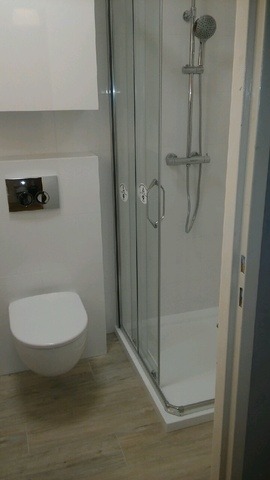 Nowoczesna łazienka po remoncie: podwieszana toaleta, kabina prysznicowa z przesuwnymi drzwiami, jasne płytki podłogowe, minimalistyczna szafka wisząca.