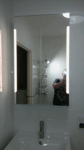 Biała umywalka z szafką lustrzaną i oświetleniem LED w łazience. W lustrze odbija się osoba robiąca zdjęcie. Widoczne białe płytki i grzejnik.