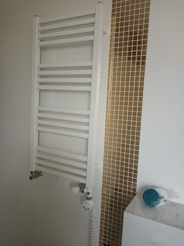 Biały, drabinkowy grzejnik łazienkowy na ścianie z mozaiką. Nowoczesny design, widoczne przyłącza i termostat. Minimalistyczne wnętrze łazienki.