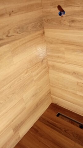 Wykończona łazienka z drewnopodobnymi panelami na ścianach i podłodze. Widoczny odpływ liniowy i wystająca rura. Ciepłe oświetlenie.