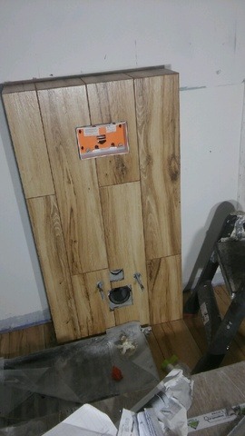 Montaż stelaża podtynkowego z obudową z płytek imitujących drewno. Widoczne otwory na przyłącza, puszka elektryczna. W tle drabina malarska i resztki materiałów budowlanych.