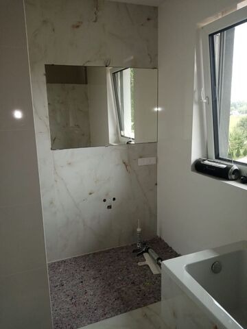 Wnętrze łazienki w trakcie prac wykończeniowych: marmurowe płytki ścienne, przygotowane miejsce na lustro, widoczne narzędzia i wanna. Okno z widokiem na zieleń.