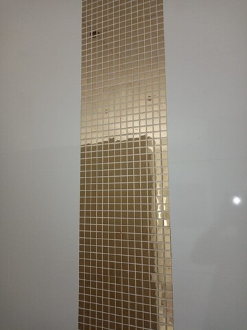 Wąski pas złotej mozaiki kwadratowej, ułożonej pionowo na gładkiej, białej ścianie, z widocznymi otworami montażowymi.