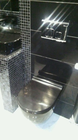 Czarna, podwieszana toaleta z deską w połysku, obok umywalka obłożona mozaiką. Nowoczesna armatura w łazience wykończonej płytkami w odcieniach czerni i srebra.