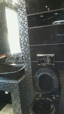 Elegancka, czarna łazienka z umywalką na mozaikowym postumencie i wiszącą toaletą. Ściany wykończone czarnym marmurem i mozaiką.