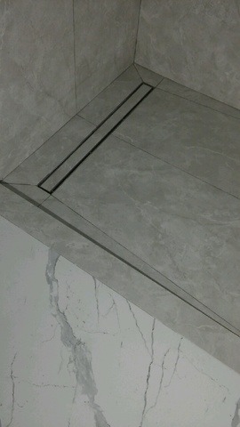 Nowoczesny odpływ liniowy w kabinie prysznicowej, wykończonej szarymi i białymi płytkami imitującymi marmur. Minimalistyczny design, precyzyjne ułożenie płytek.