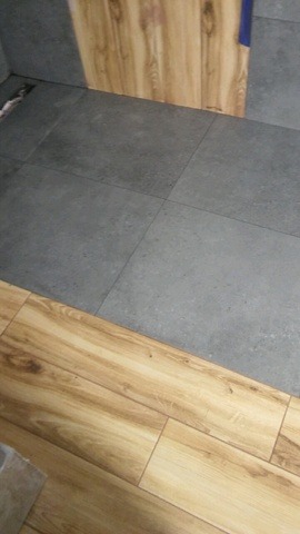 Ułożone szare płytki podłogowe graniczące z panelami imitującymi drewno. Widoczny fragment ściany z drewnopodobną okładziną.