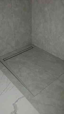 Wykończona strefa prysznicowa z szarymi płytkami imitującymi kamień i liniowym odpływem. Uwagę przykuwa kontrast między szarością kabiny a marmurkowym wzorem podłogi.