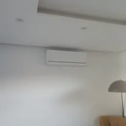 Biała klimatyzacja ścienna zamontowana w jasnym wnętrzu. Widoczne oświetlenie sufitowe i fragment lampy podłogowej. Minimalistyczny design.