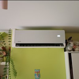 MR-KLIMA SYSTEM Robert Duszyński - Szary klimatyzator Rotenso na zielonej ścianie w pokoju, obok rośliny doniczkowe i pluszaki. Widoczny wyświetlacz z temperaturą 22 stopnie.