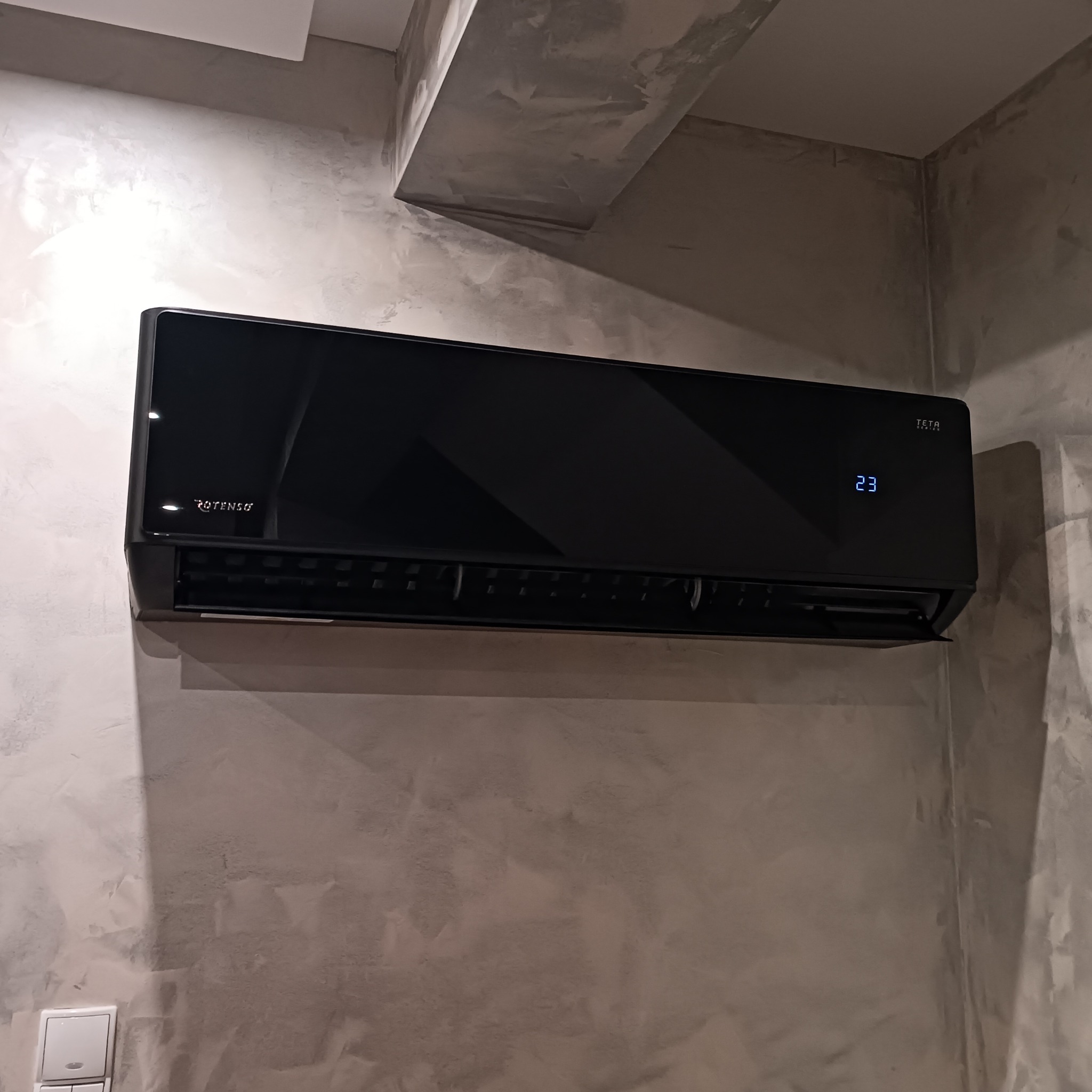 Czarna klimatyzacja Rotenso Teta Series na szarej ścianie z efektem betonu. Widoczny wyświetlacz z temperaturą 23 stopnie Celsjusza. Nowoczesny design.