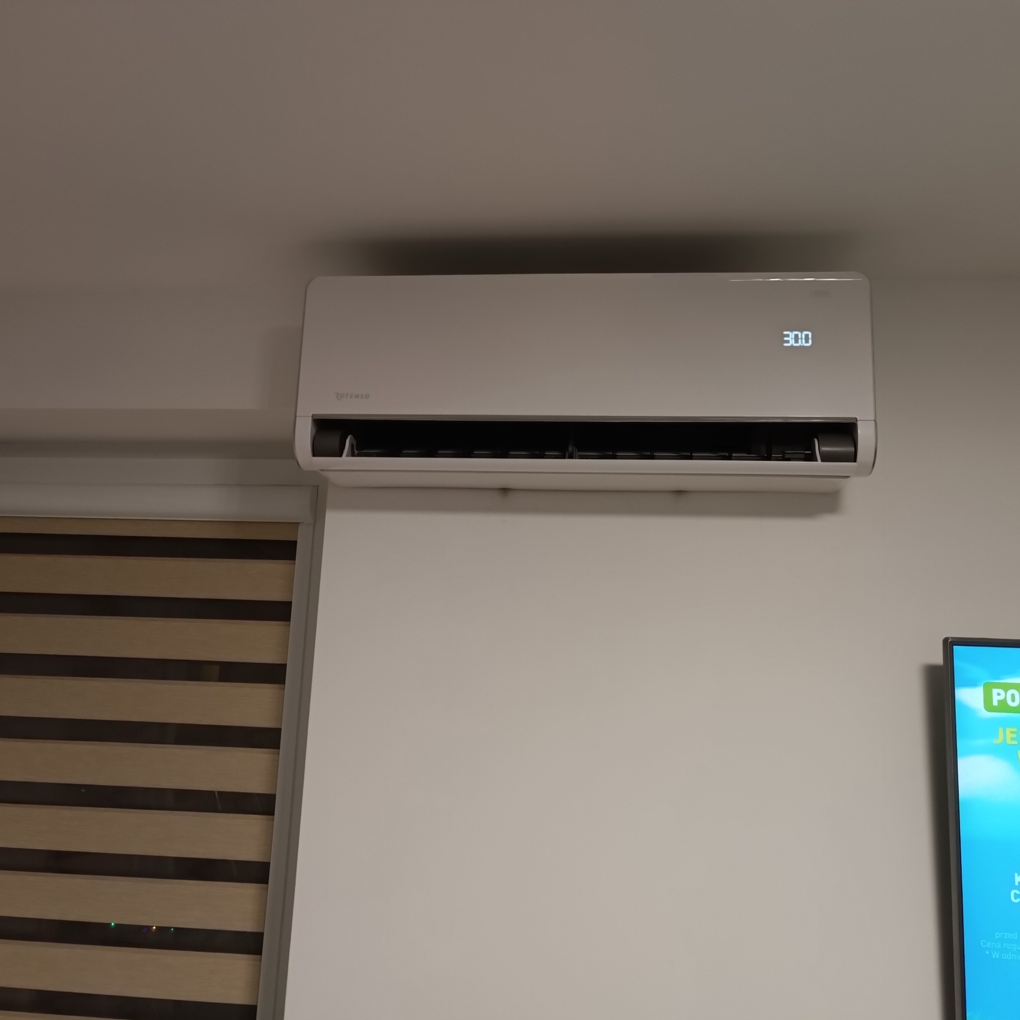 Biały klimatyzator Rotenso na ścianie, wyświetlona temperatura 30 stopni Celsjusza, obok okno z żaluzjami i fragment telewizora.