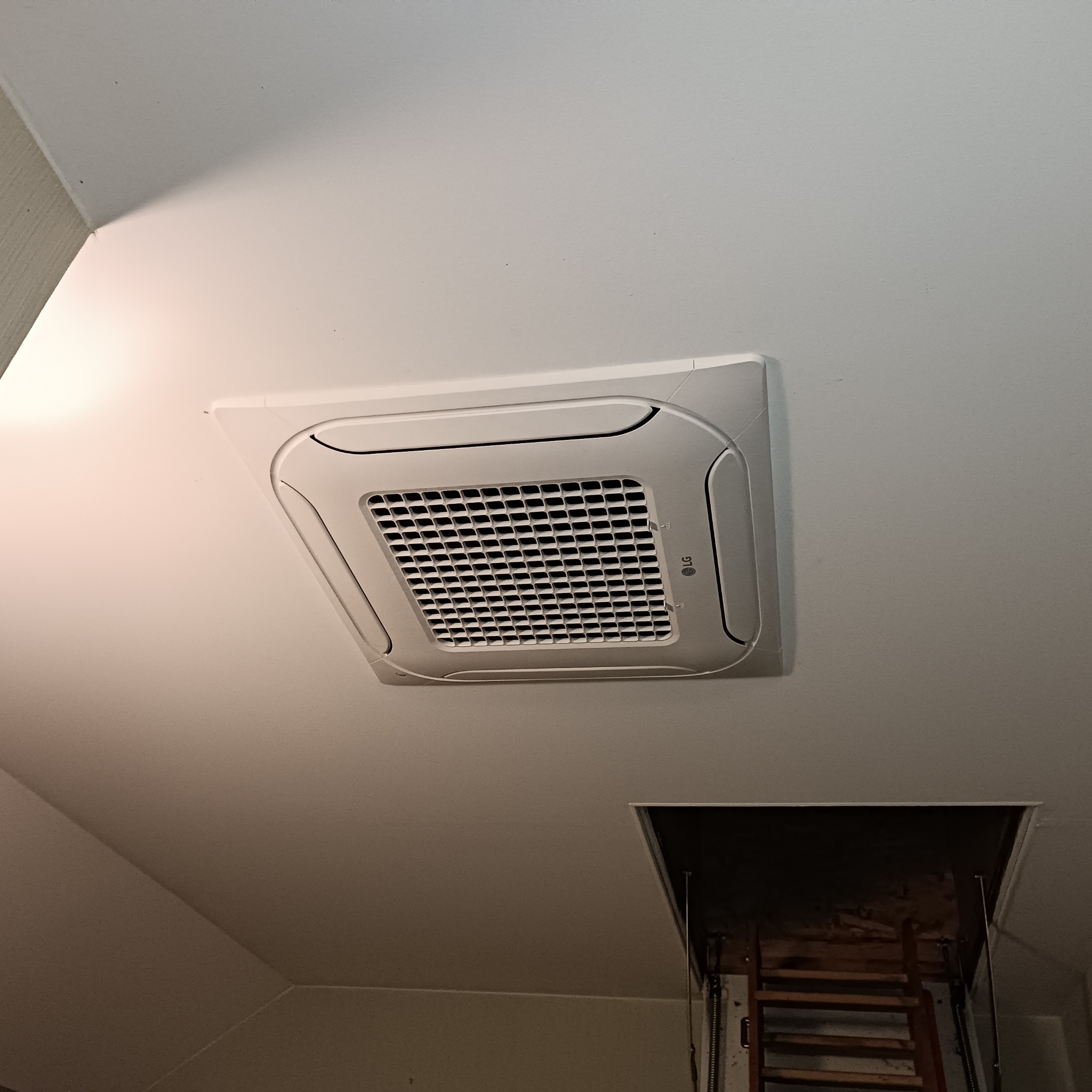 Kasetonowa klimatyzacja LG zamontowana na suficie poddasza, widoczny otwór włazu strychowego z rozkładaną drabinką. Ujęcie z dołu, oświetlenie górne.