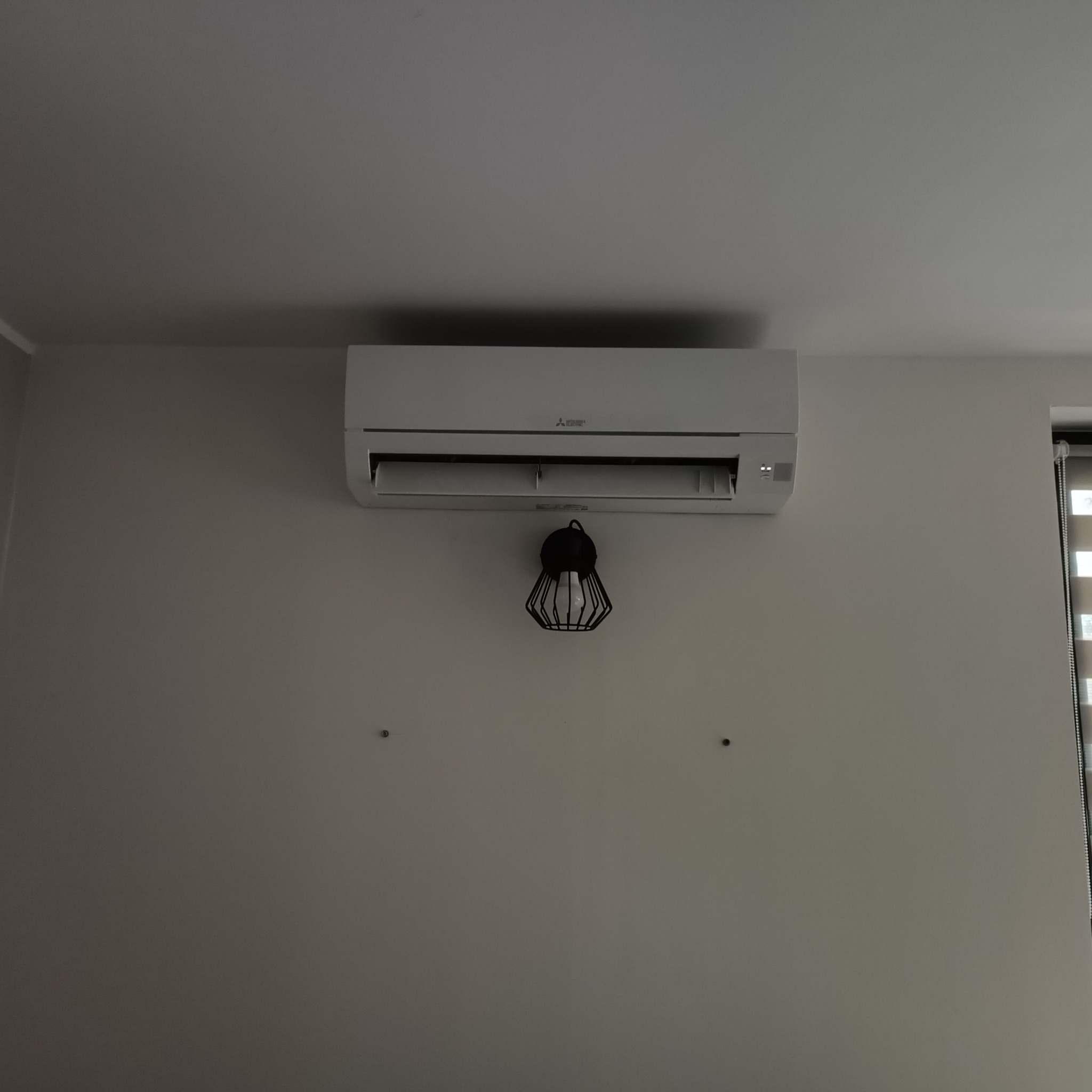 Klimatyzator Mitsubishi na ścianie w jasnym pomieszczeniu. Pod nim wisi dekoracyjna lampa. Widoczne rolety w oknie po prawej stronie.