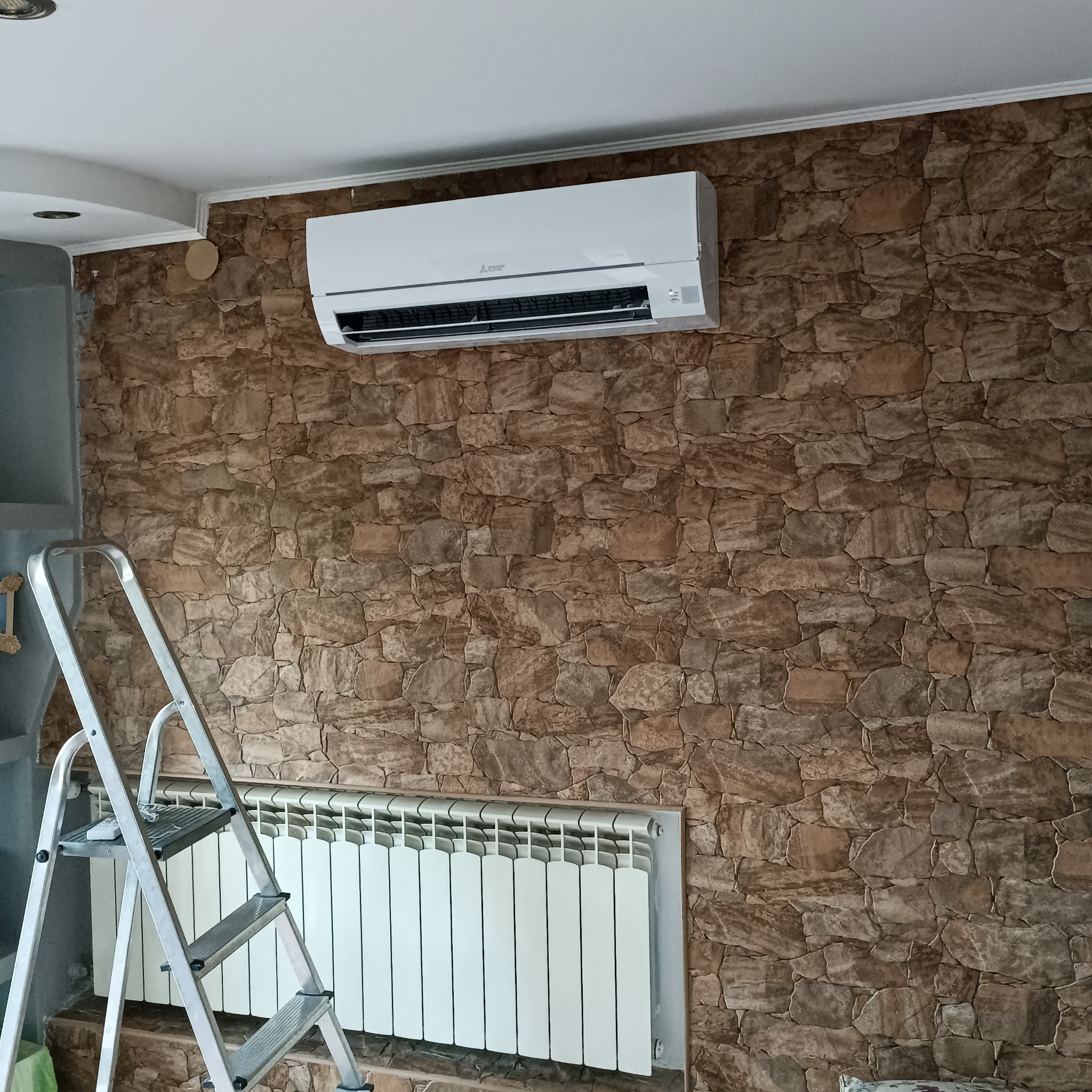 Biały klimatyzator Mitsubishi zamontowany na ścianie z kamienną tapetą. Pod nim biały grzejnik. Z lewej drabina, sugerująca niedawny montaż.