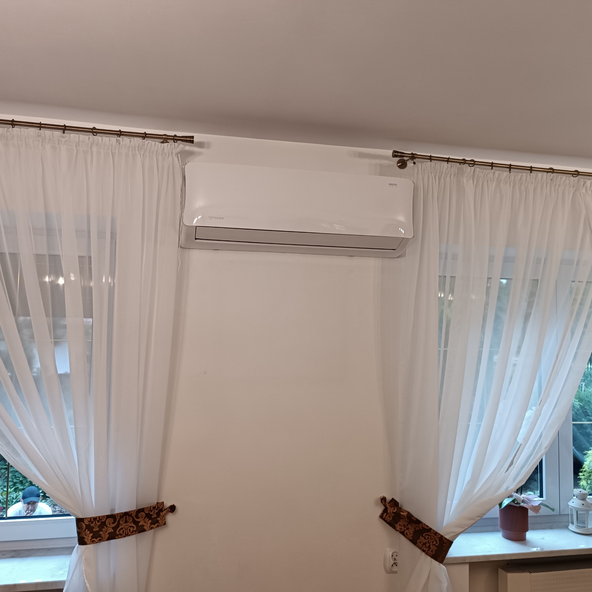 Biały klimatyzator ścienny zamontowany w pokoju z oknami zasłoniętymi białymi firanami, z dekoracyjnymi upięciami po bokach. Widoczne gniazdko elektryczne.