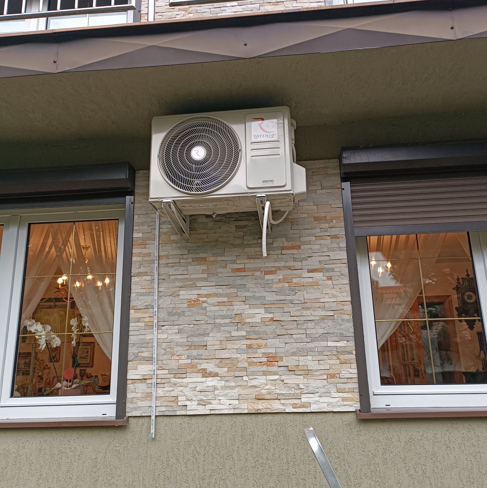 Biały klimatyzator Rotenso zamontowany na elewacji budynku z dekoracyjnej cegły, widoczny pomiędzy dwoma oknami z roletami zewnętrznymi.