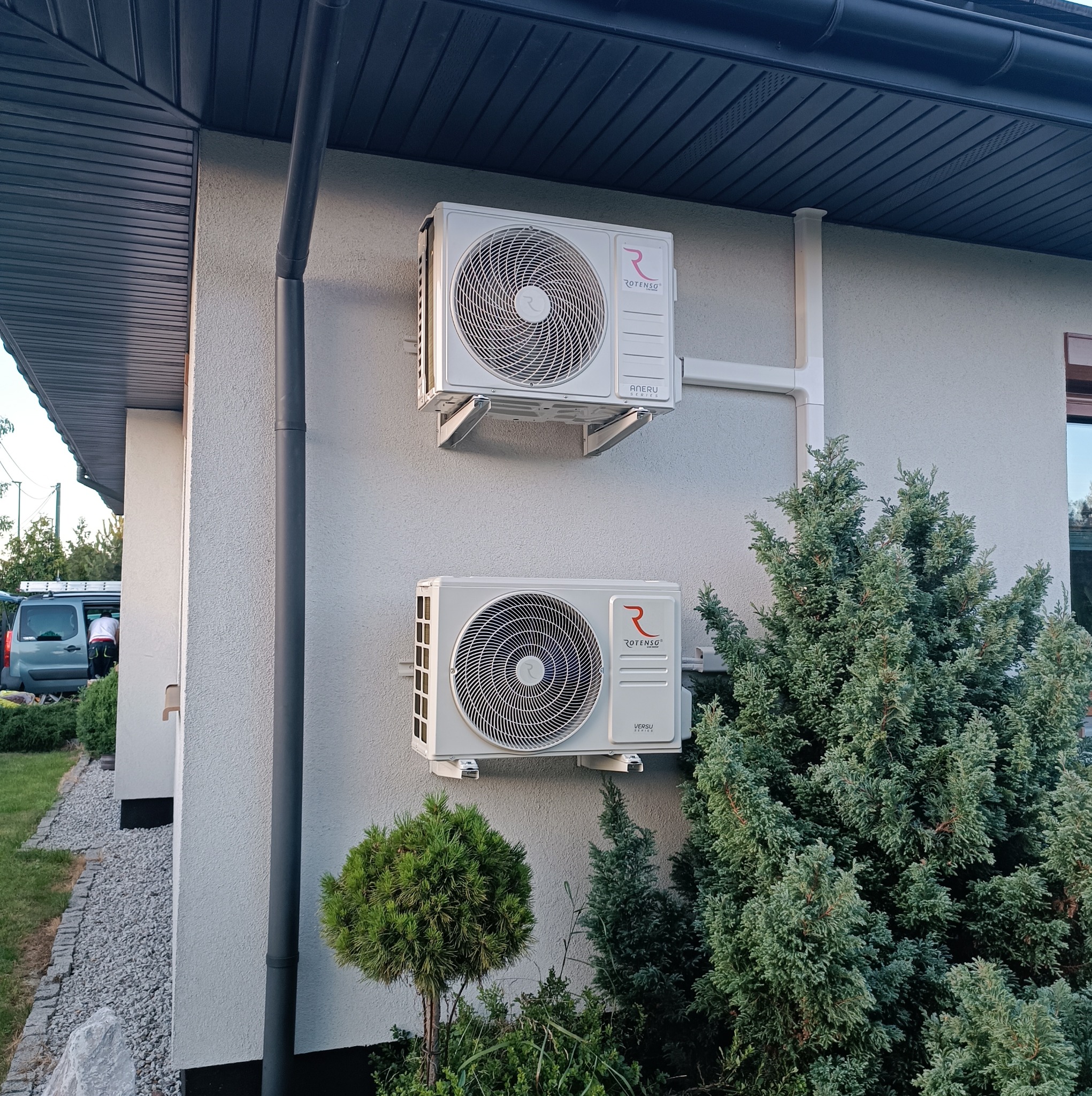 Dwa białe klimatyzatory Rotenso zamontowane na elewacji domu. Widoczne rury odprowadzające, zieleń wokół i fragment dachu.