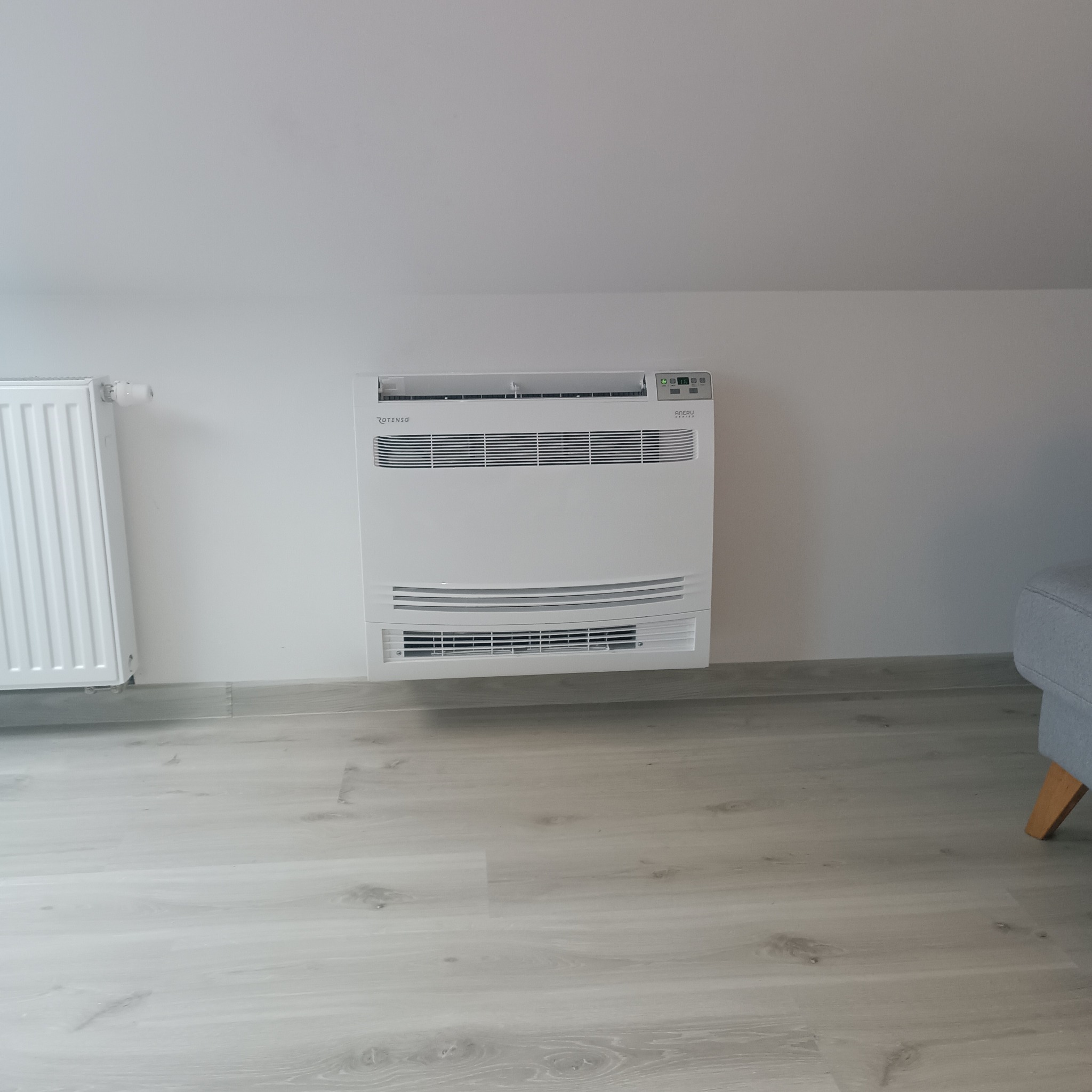 Biała klimatyzacja podłogowa Rotenso Aneri w jasnym wnętrzu z grzejnikiem i fragmentem szarego fotela. Minimalistyczny design, nowoczesne rozwiązanie grzewczo-chłodzące.