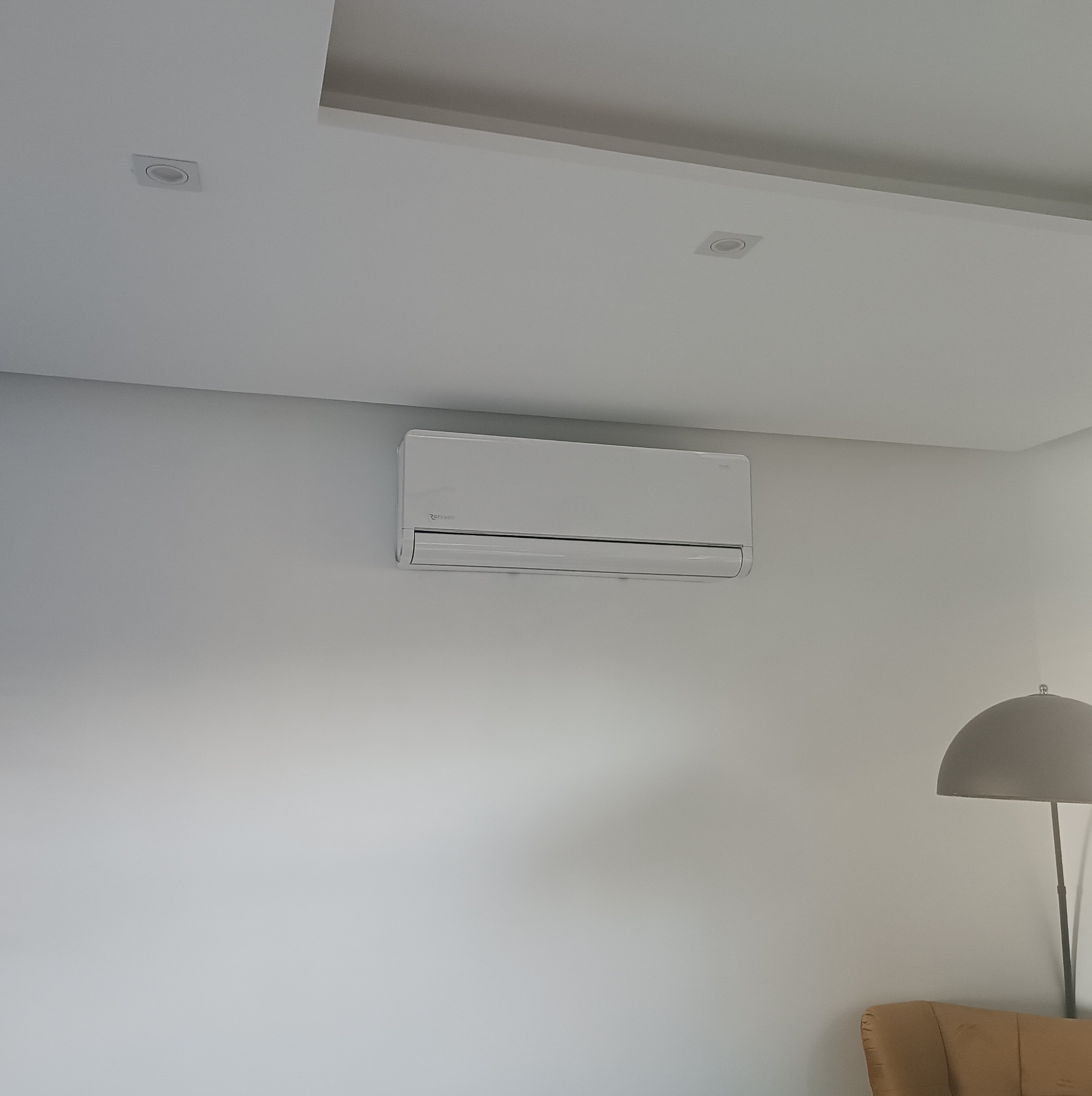 Biała klimatyzacja ścienna zamontowana w jasnym wnętrzu. Widoczne oświetlenie sufitowe i fragment lampy podłogowej. Minimalistyczny design.