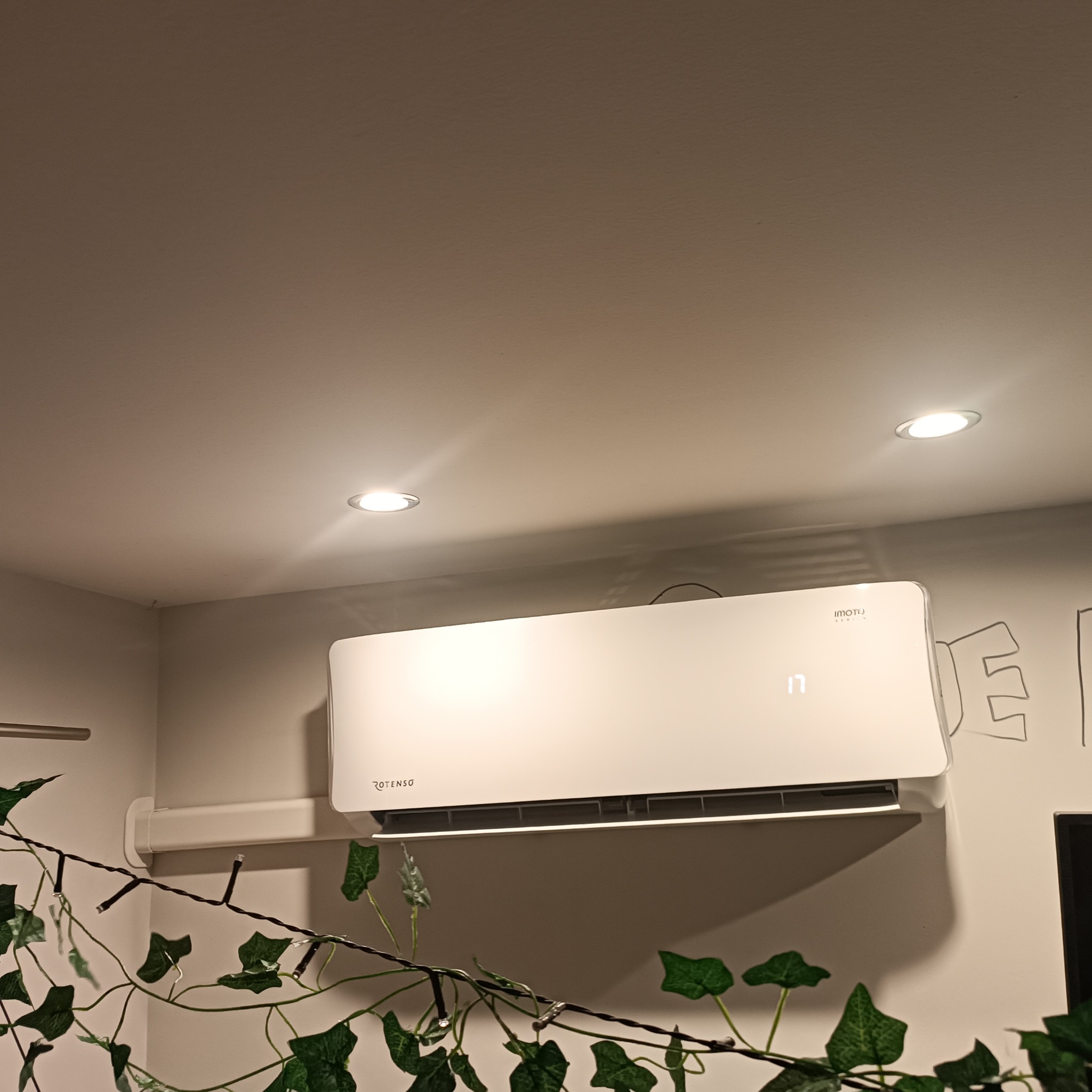 Biała klimatyzacja Rotenso zamontowana na ścianie, widoczna dekoracja z bluszczu i oświetlenie sufitowe. Kąt ujęcia z dołu.