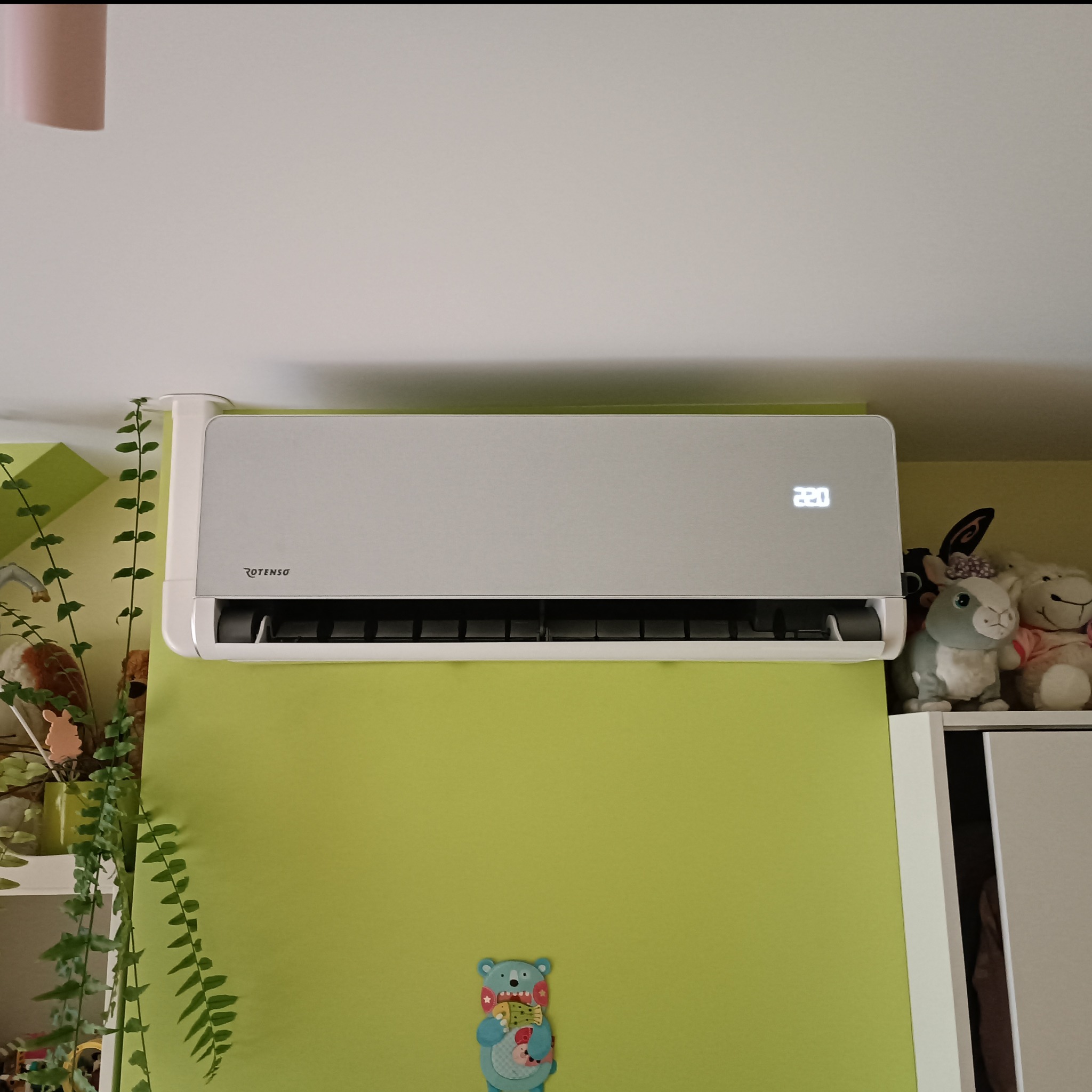 Szary klimatyzator Rotenso na zielonej ścianie w pokoju, obok rośliny doniczkowe i pluszaki. Widoczny wyświetlacz z temperaturą 22 stopnie.