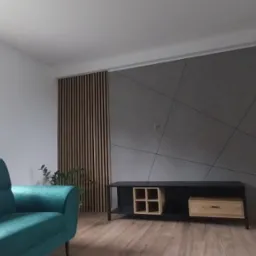 Szary panel ścienny z geometrycznym wzorem, drewniane lamele i szafka RTV w nowoczesnym salonie z zieloną sofą. Okno z widokiem na zieleń.