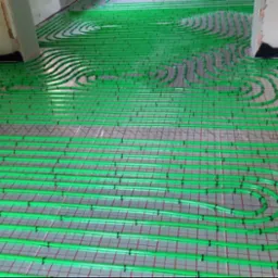 Zielone rury ogrzewania podłogowego ułożone na siatce, gotowe do zalania posadzką, widoczne fragmenty ścian.