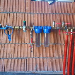 Usługi hydrauliczno-remontowe Robert Łebek - Instalacja wodna z filtrami i zaworami na tle ściany z cegły dziurawki. Widoczne czerwone rury z opisami, manometr i niebieski klucz do filtrów.