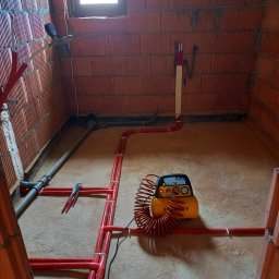 Usługi hydrauliczno-remontowe Robert Łebek - Czerwone rury instalacji ogrzewania podłogowego w surowym wnętrzu z ceglanymi ścianami. Kompresor Stanley i szczypce obok. Widok z góry na podłogę.