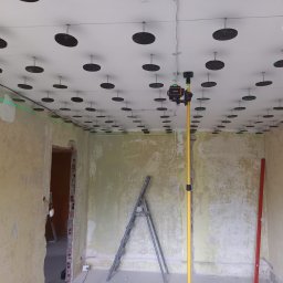 Usługi hydrauliczno-remontowe Robert Łebek - Montaż sufitu podwieszanego z użyciem lasera krzyżowego. Widoczne profile i elementy mocujące na suficie. Ściany w stanie surowym, drabina oparta o ścianę.