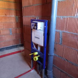 Usługi hydrauliczno-remontowe Robert Łebek - Stelaż podtynkowy Geberit Duofix zamontowany przy czerwonej ścianie z pustaków. Widoczne rury kanalizacyjne i dopływowe. Instalacja w trakcie montażu.