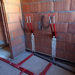 Usługi hydrauliczno-remontowe Robert Łebek - Czerwone rury instalacji wodnej zamocowane do ściany z cegły, zabezpieczone pianką montażową. Szare rury kanalizacyjne wzdłuż podłogi.
