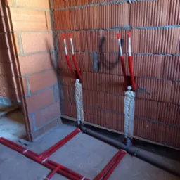 Czerwone rury instalacji wodnej zamocowane do ściany z cegły, zabezpieczone pianką montażową. Szare rury kanalizacyjne wzdłuż podłogi.