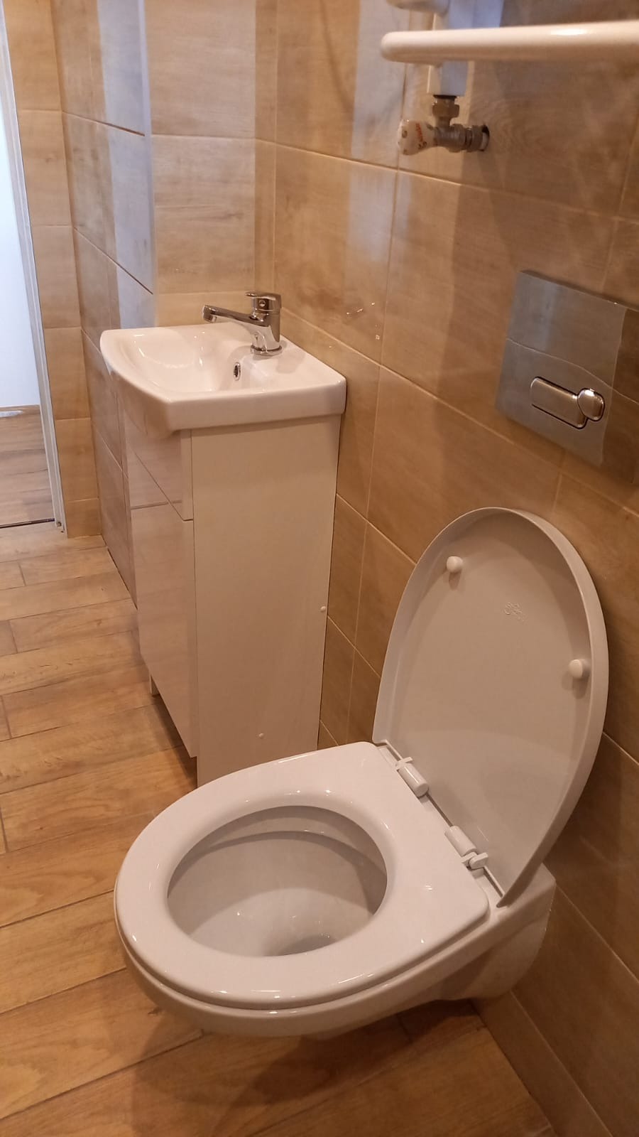 Jasna łazienka po remoncie: biała umywalka z szafką, toaleta z otwartą klapą i beżowe płytki imitujące drewno. Widoczny grzejnik drabinkowy.