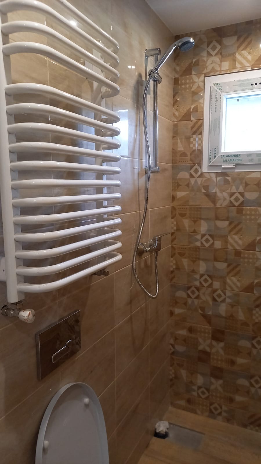 Wykończona łazienka z beżowymi płytkami, grzejnikiem drabinkowym, prysznicem i oknem. Widoczne detale armatury i wzór płytek.