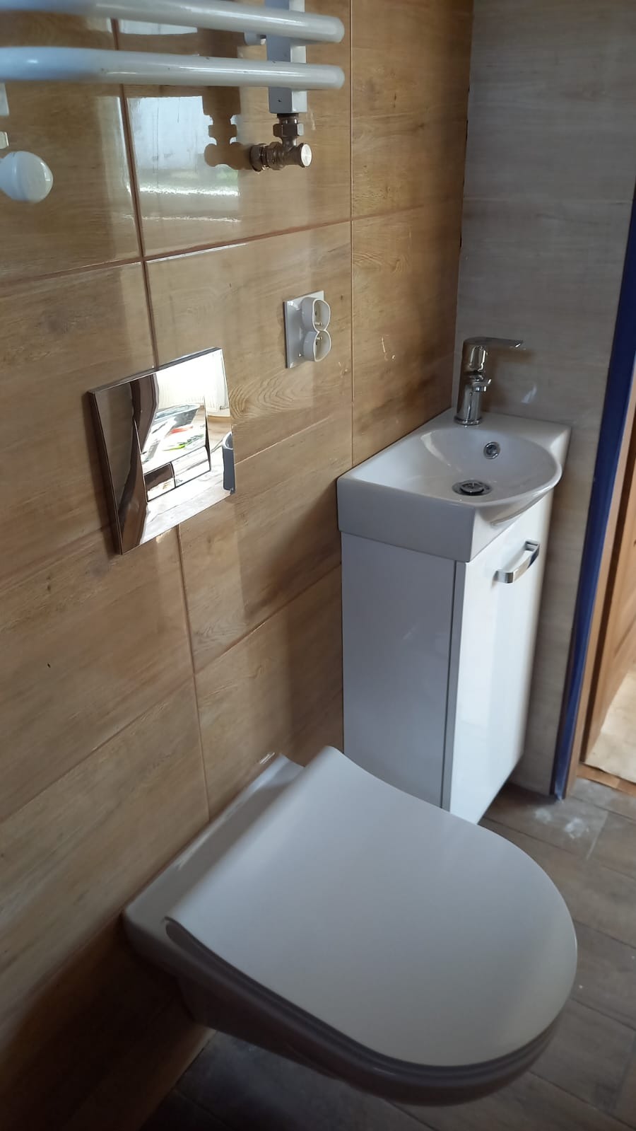 Nowoczesna toaleta podwieszana z umywalką narożną i grzejnikiem drabinkowym na ścianie z płytek imitujących drewno. Widoczne elementy instalacji.