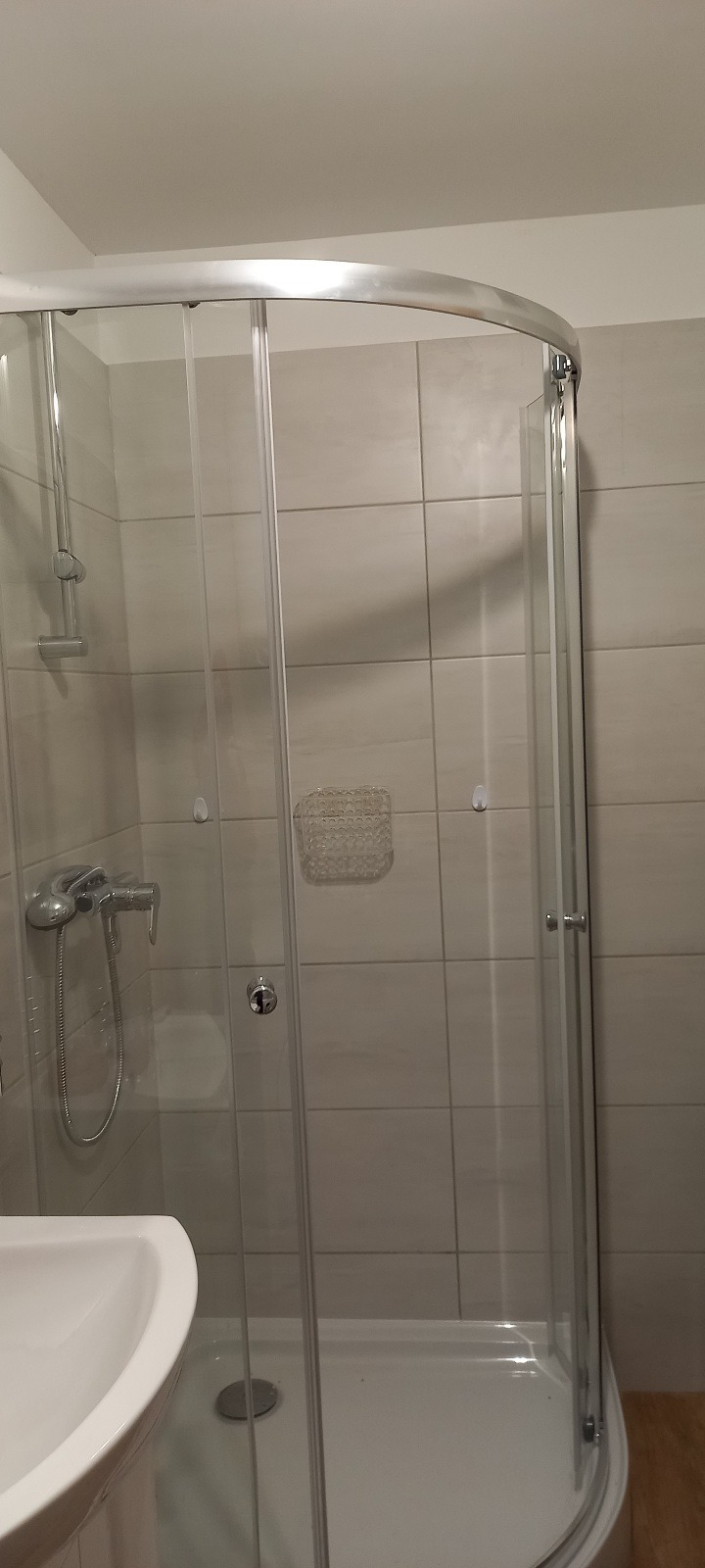 Nowoczesna kabina prysznicowa z chromowanymi profilami i szklanymi drzwiami, umywalka z baterią w tle, jasne płytki na ścianach, minimalistyczny design.