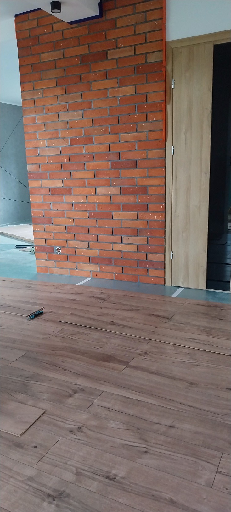 Montaż paneli podłogowych w pokoju z dekoracyjną ścianą z cegły i jasnymi drzwiami. Widoczne narzędzia pracy i fragmenty nieułożonych paneli.