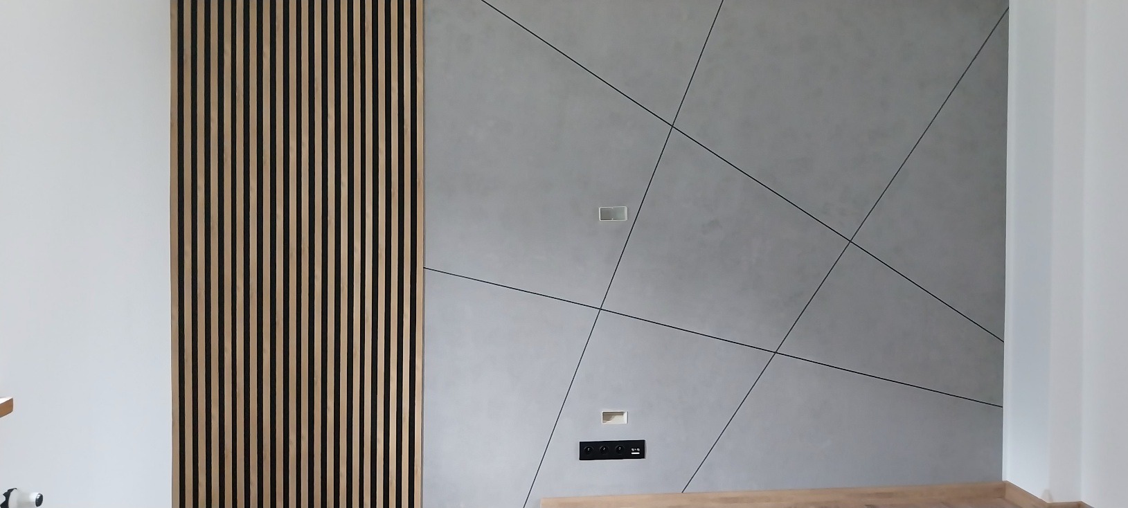 Nowoczesna ściana z dekoracyjnymi panelami lamelowymi i płytami betonowymi z czarnymi liniami podziału, gniazdka elektryczne.