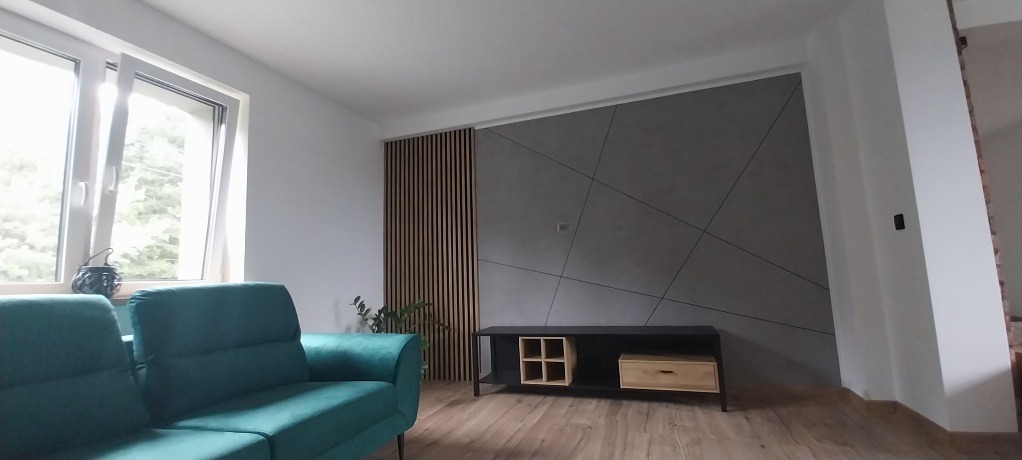 Szary panel ścienny z geometrycznym wzorem, drewniane lamele i szafka RTV w nowoczesnym salonie z zieloną sofą. Okno z widokiem na zieleń.