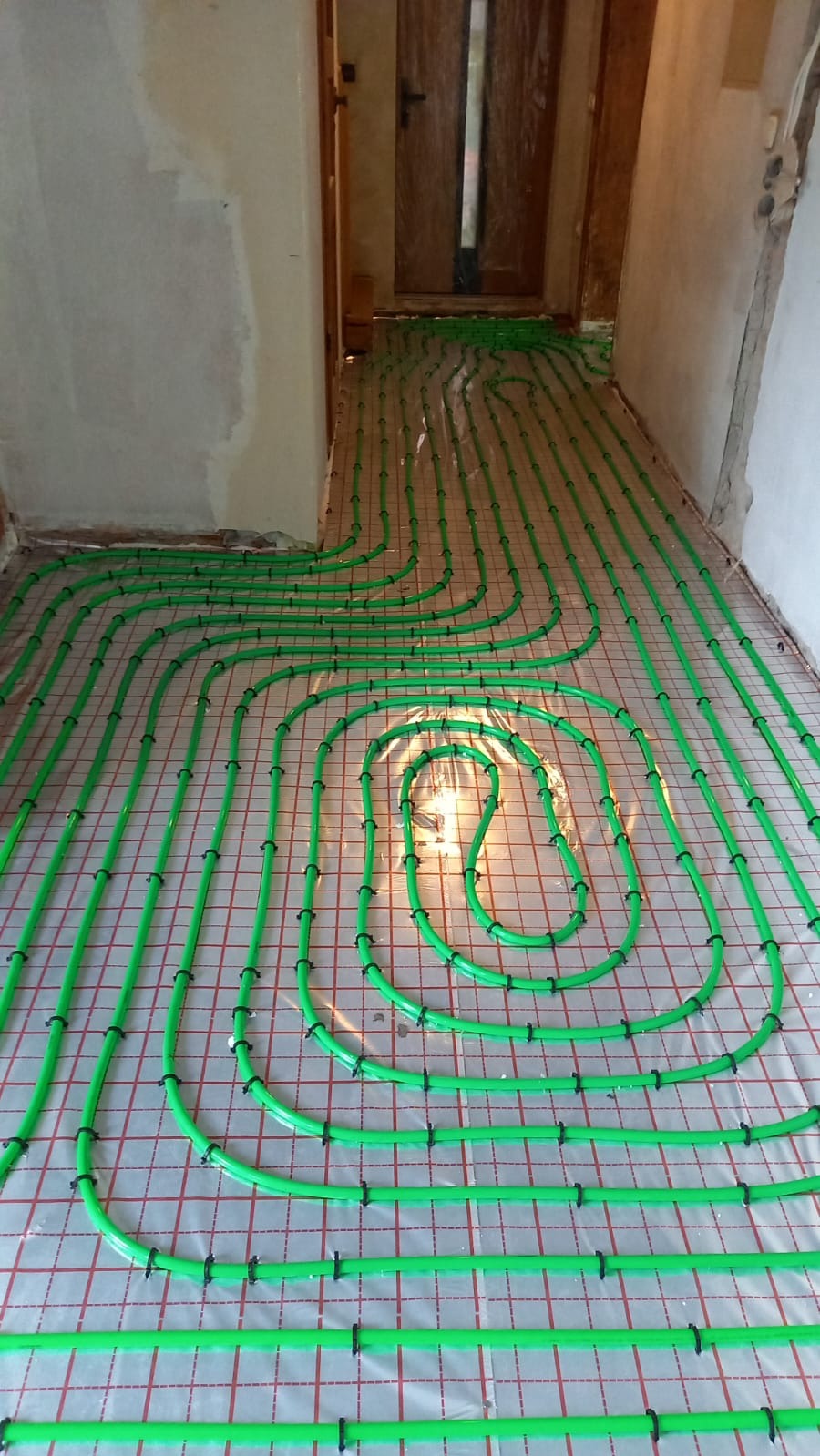 Zielone rury ogrzewania podłogowego ułożone na folii z siatką w surowym wnętrzu, widoczne elementy mocujące i fragment drzwi w tle.