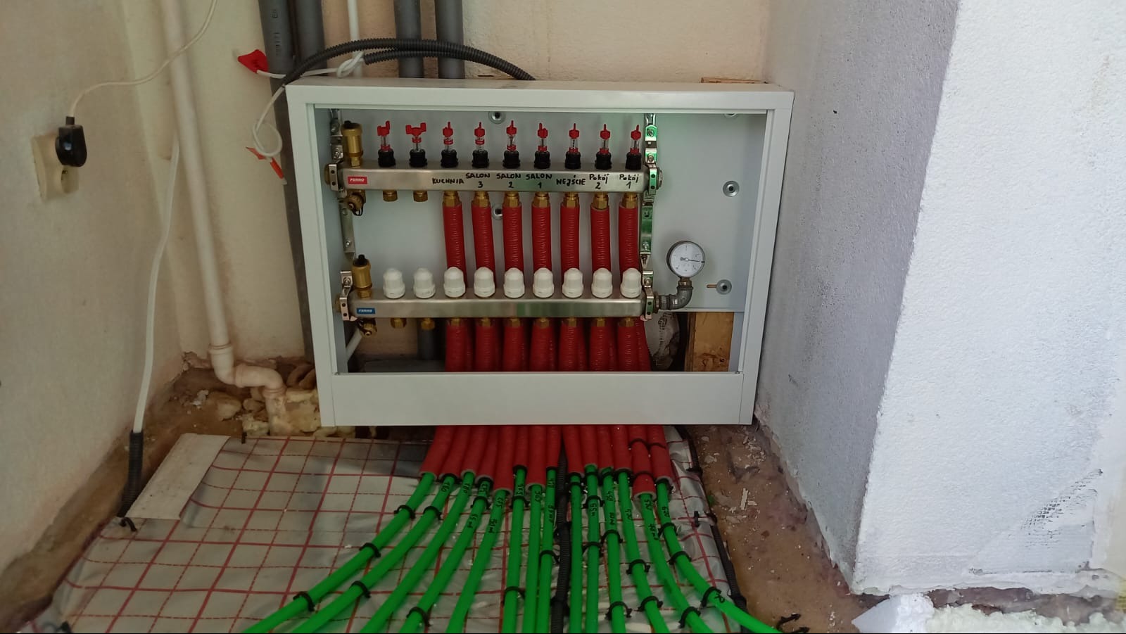 Rozdzielacz ogrzewania podłogowego z rurami w izolacji, zamontowany na ścianie, widok na ułożoną matę grzewczą. Elementy instalacji opisane.