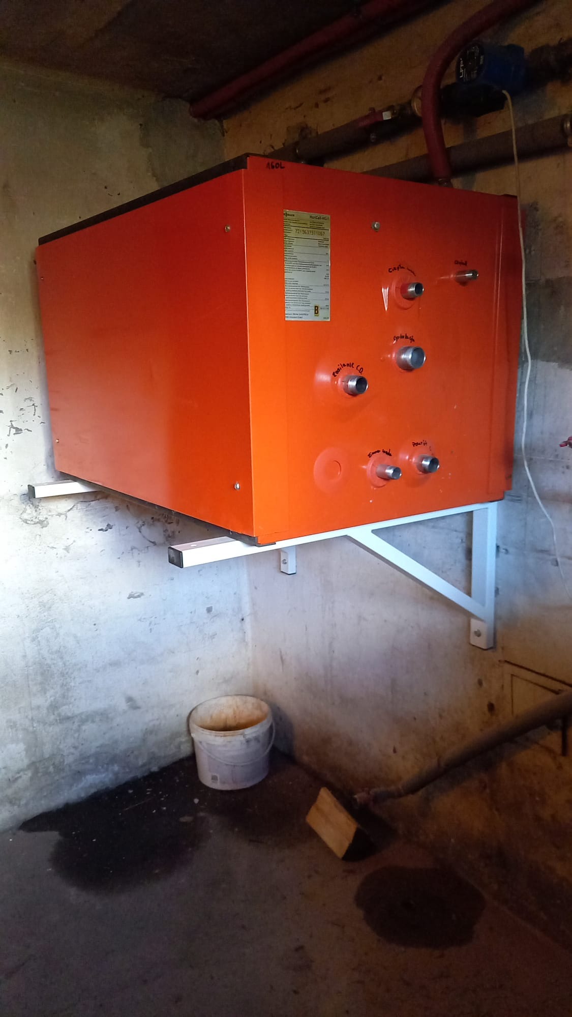 Pomarańczowy piec CO zamontowany na wspornikach w surowym pomieszczeniu. Widoczne przyłącza i etykieta informacyjna. U dołu wiadro i kawałek drewna.